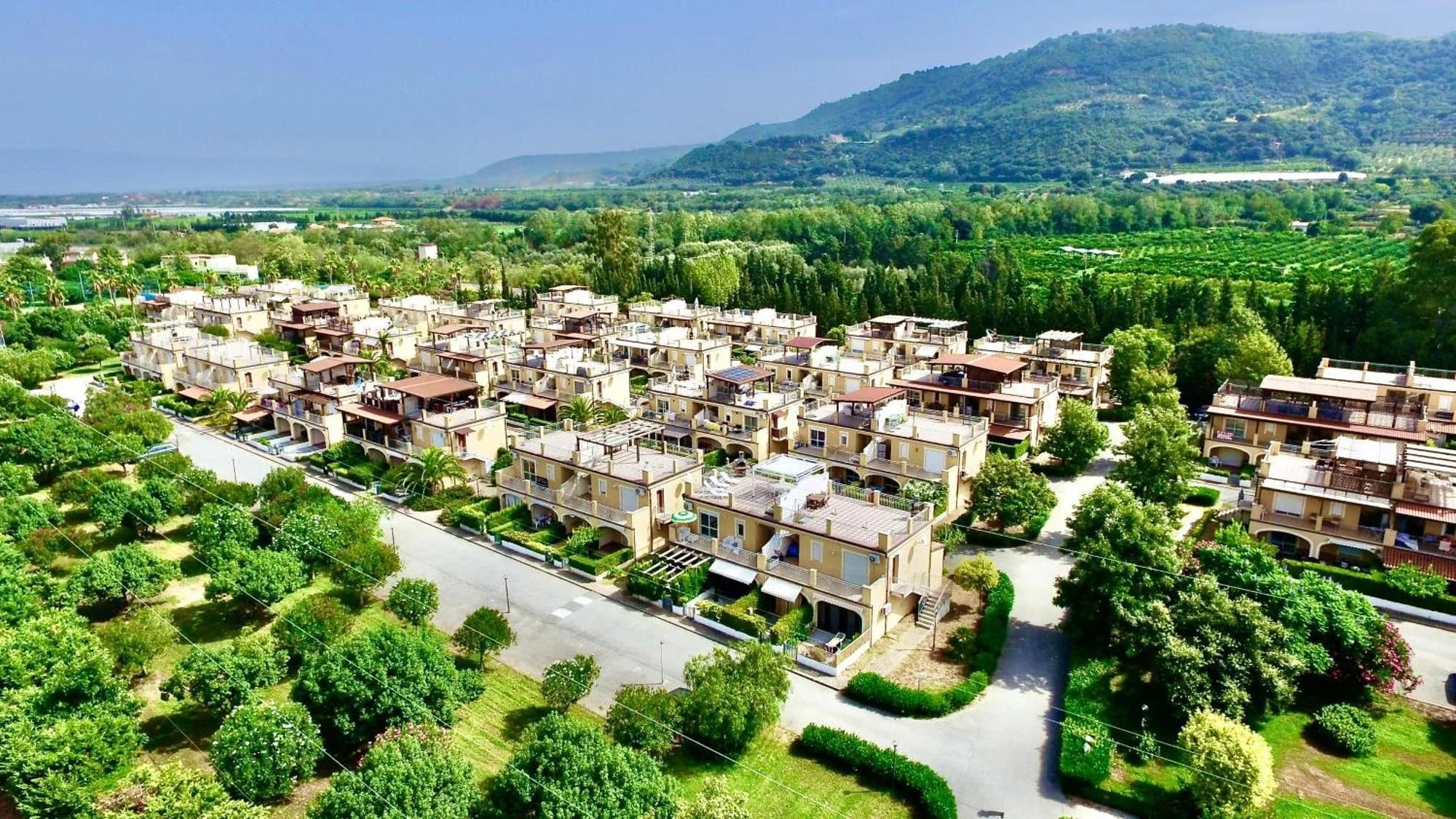 Veduta aerea del Pizzo Beach Club: palazzine con terrazze tra viali alberati e giardini, con le colline sullo sfondo